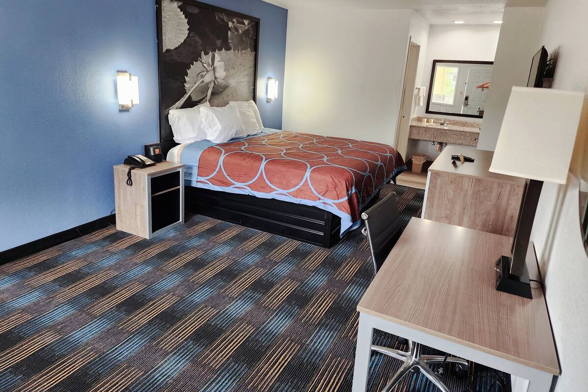 Una habitación de hotel con cama, mesita de noche, escritorio, silla y un cuadro grande en la pared.