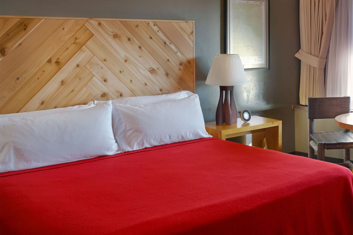 A hotel room with a large bed, a wooden headboard, a bedside table with a lamp, and a chair.