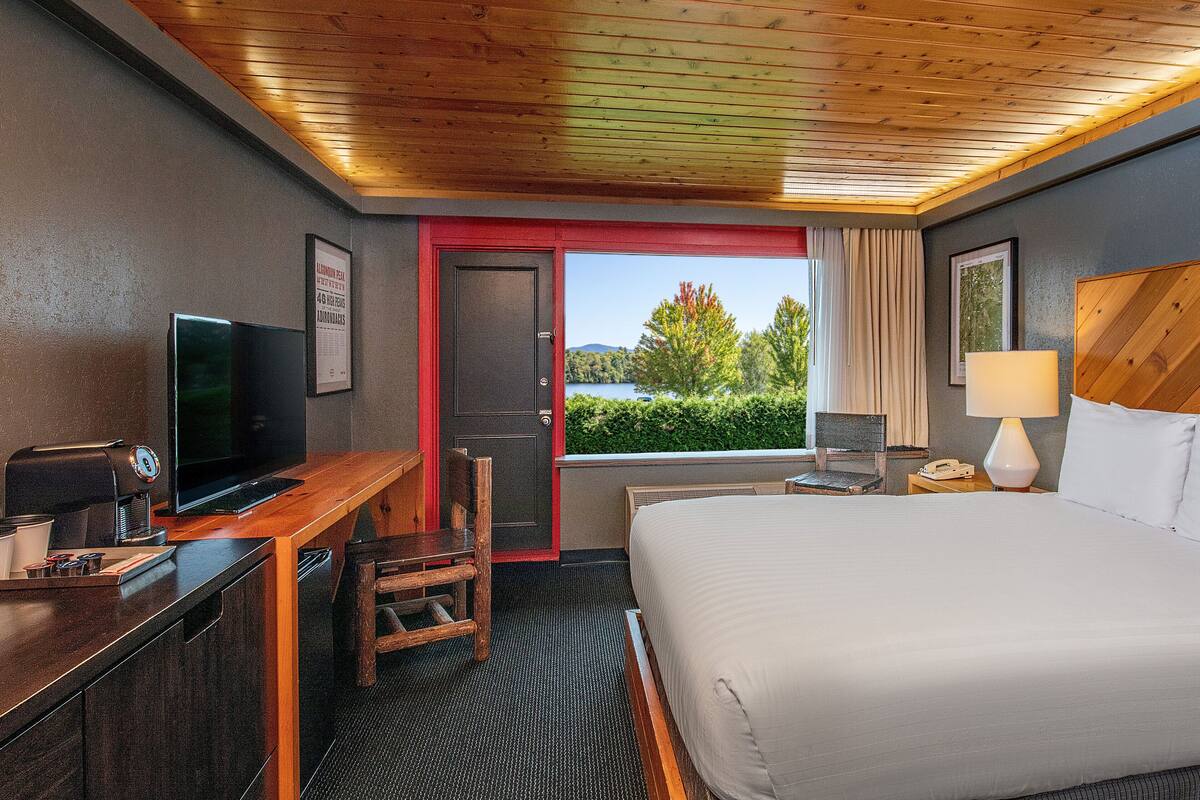 A hotel room with a bed, a desk with a TV, a chair, a window with a view of trees and water, and a wooden ceiling.