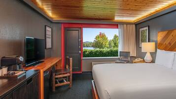 A hotel room with a bed, a desk with a TV, a chair, a window with a view of trees and water, and a wooden ceiling.