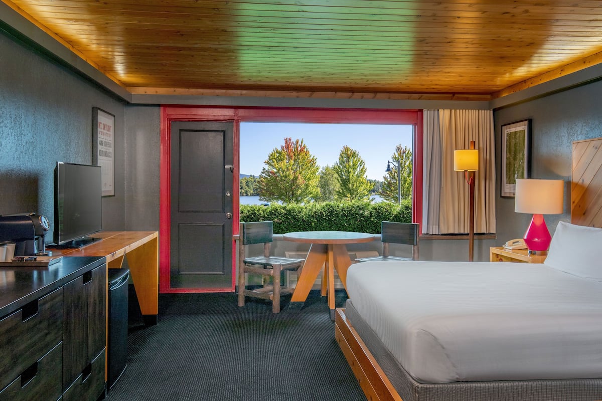 A hotel room with a bed, desk, chair, table, and a view of trees and a lake.