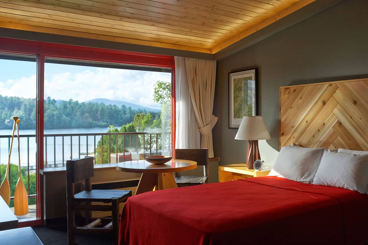A hotel room with a bed, a table with a bowl, a chair, a wooden headboard, a lamp, a painting, and a view of a lake and mountains.