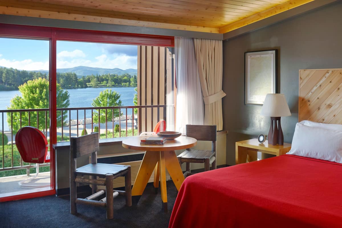 A hotel room with a bed, a table with chairs, a view of a lake, and a balcony with a chair.