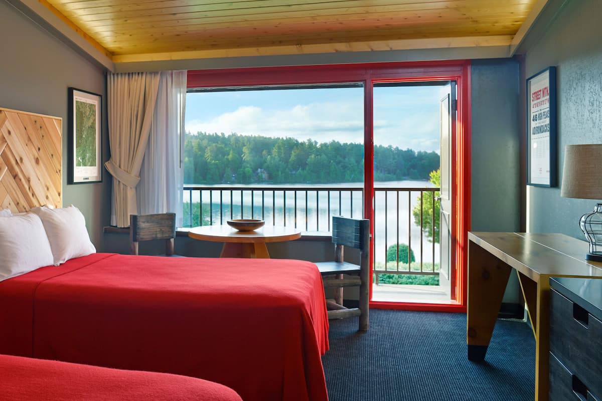 A hotel room with two beds, a desk, a chair, a table, and a balcony with a view of the lake.