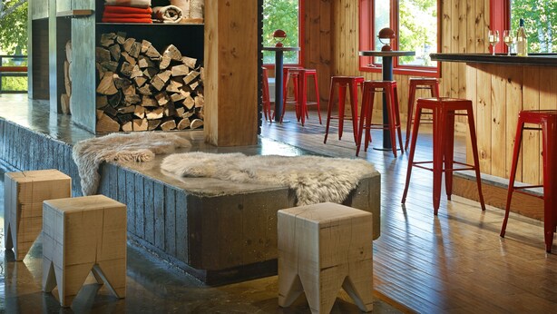 Un intérieur rustique en bois, avec une cheminée, une pile de bois de chauffage et des tabourets de bar rouges.