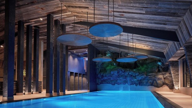 A modern indoor swimming pool with a blue-lit water surface, wooden ceiling beams, and hanging circular light fixtures. - Thermen Bad Nieuweschans (Bad Nieuweschans)