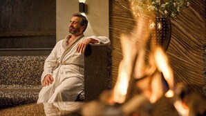 A person in a white bathrobe sitting on a bench in a spa-like setting with a fire and decorative flowers in the background. - Thermen Bad Nieuweschans (Bad Nieuweschans)