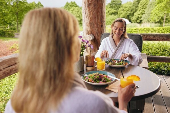 Dos personas cenando al aire libre en una mesa de madera, con una ensalada y zumo de naranja.