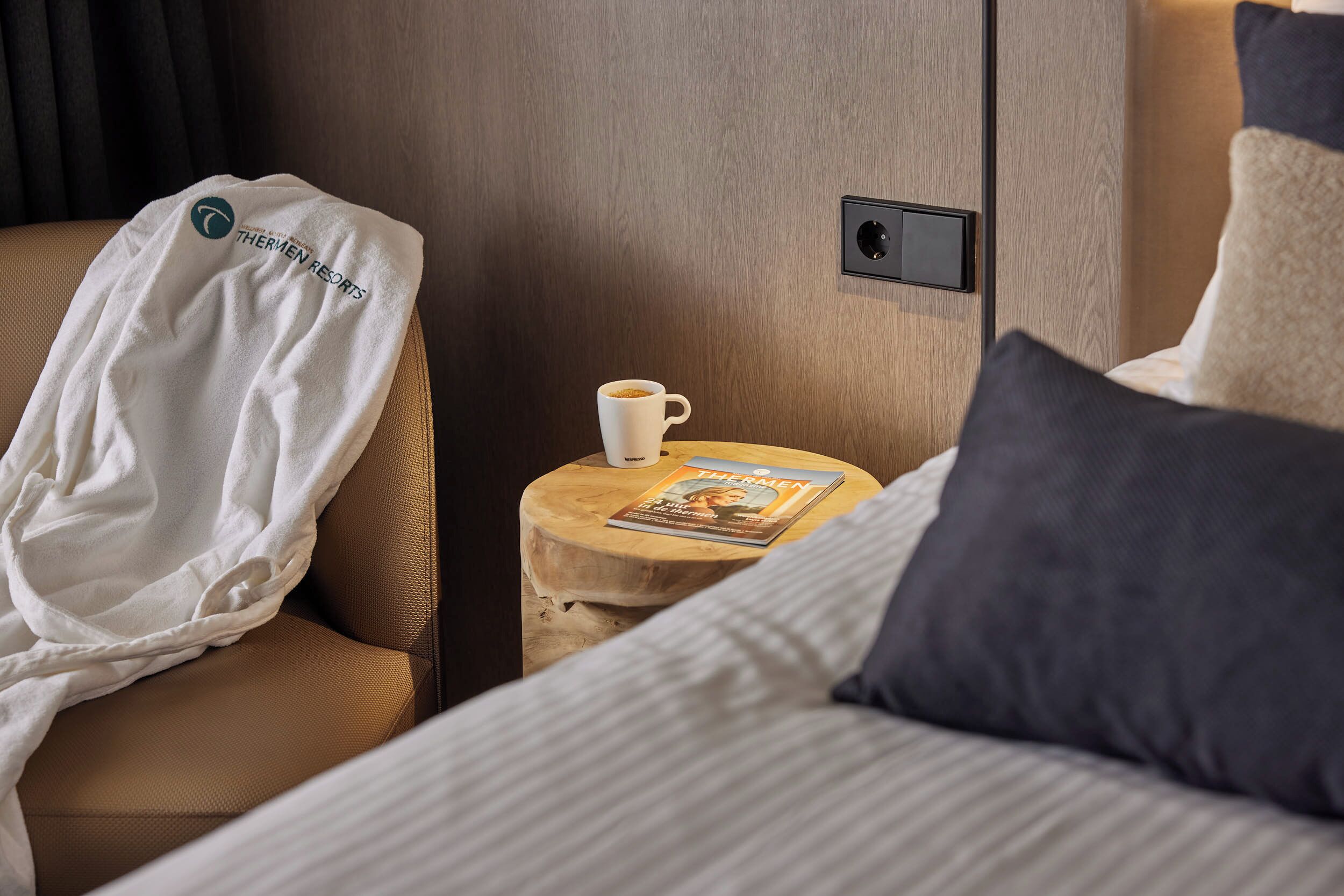 Una habitación de hotel con una cama, una mesita de noche de madera con una revista, una bata blanca y una taza de café.