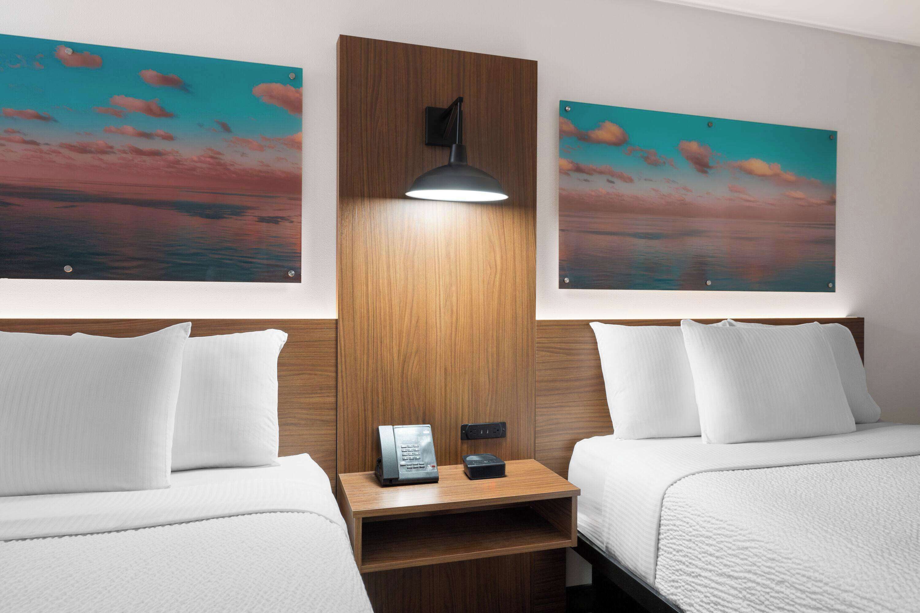 Habitación de hotel con dos camas, cabecero de madera, mesita de noche con lámpara y dos cuadros de un atardecer.