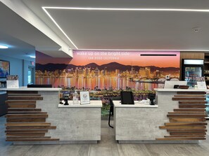 Zona de recepción de un hotel con un mural de la ciudad, un cartel de 'No Fumar' y un refrigerador lleno de bebidas.