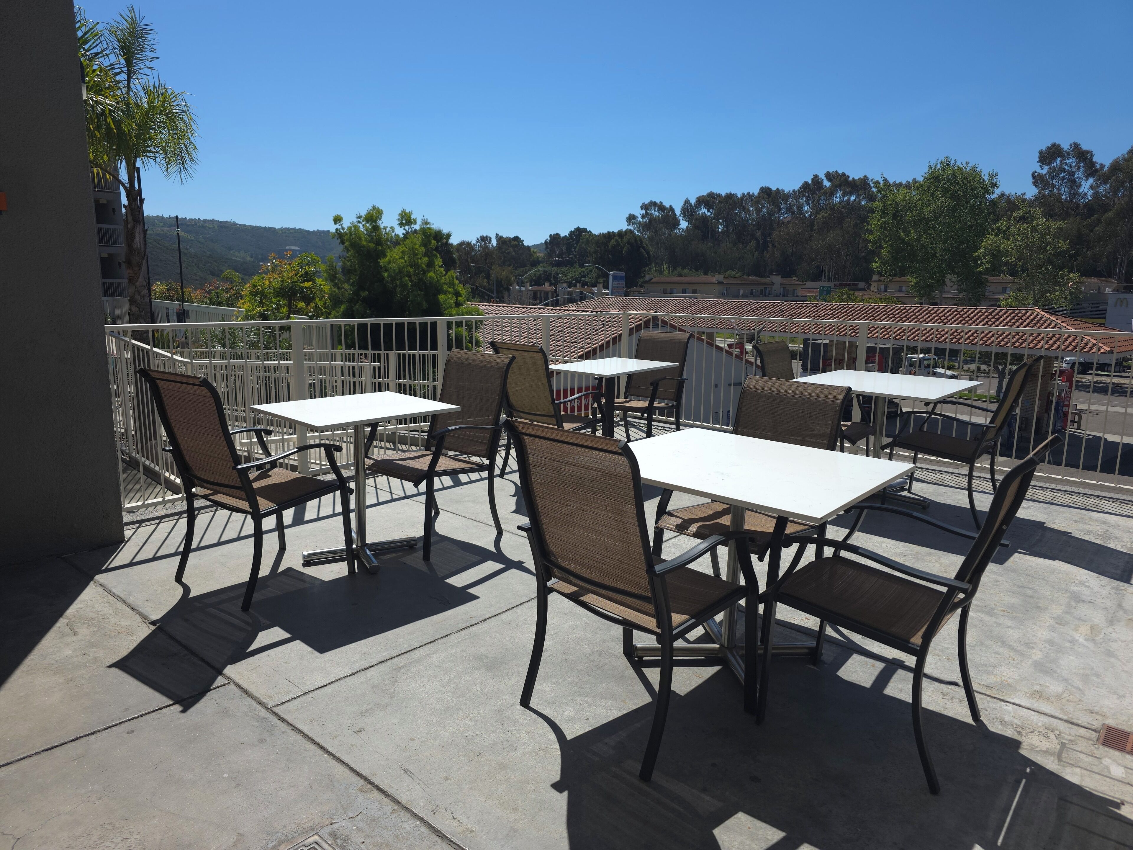An outdoor patio with tables and chairs, a white railing, and a view of a hilly landscape.