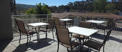 An outdoor patio with tables and chairs, a white railing, and a view of a hilly landscape.