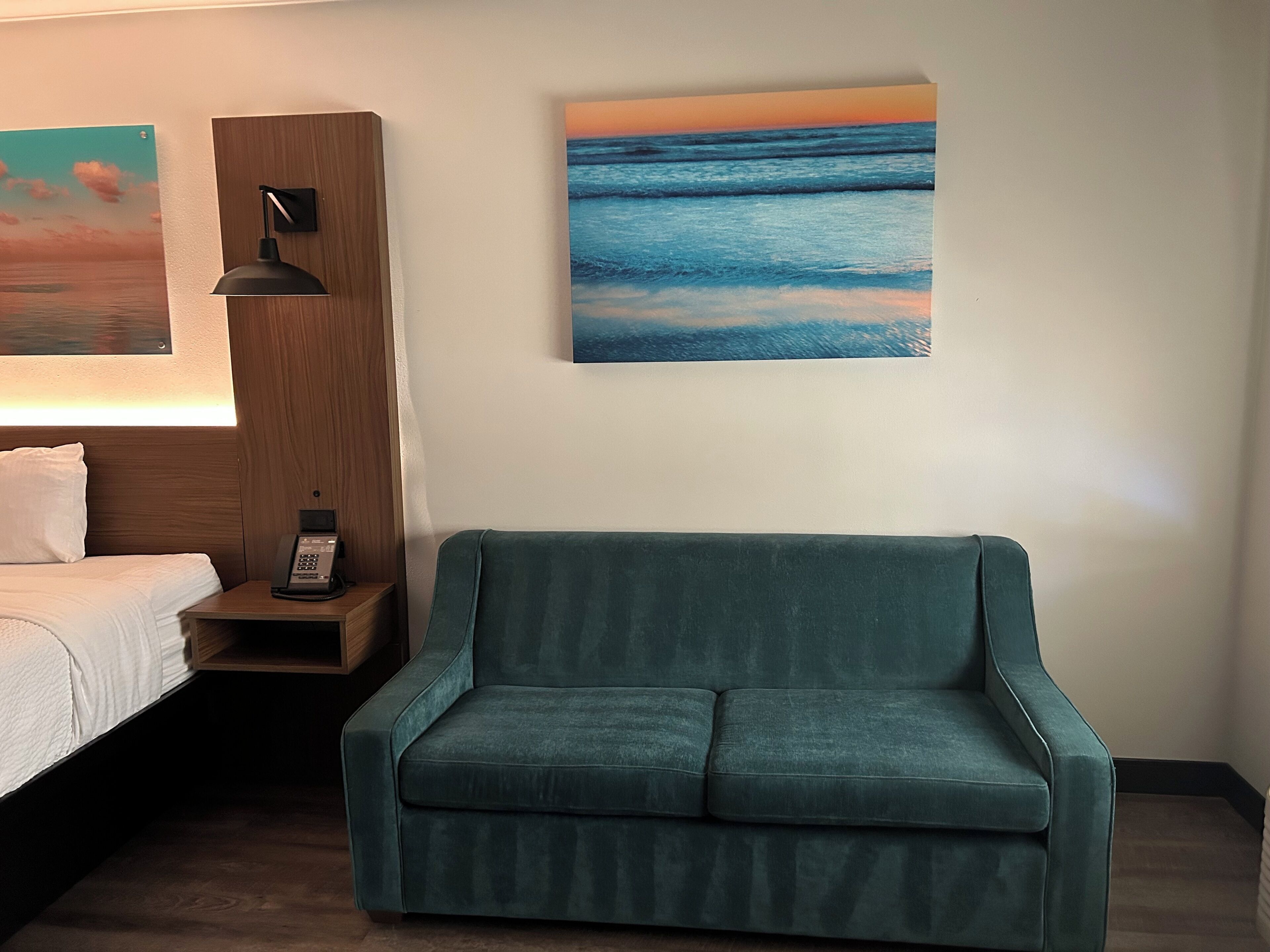 Ett hotellrum med en säng, en turkosfärgad soffa, ett nattduksbord med en lampa och två tavlor på väggen.