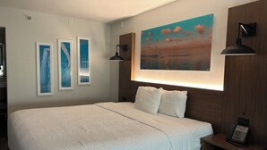 Una cama bien hecha con sábanas blancas, un cuadro colgado en la pared que muestra un atardecer y una mesita de noche con un teléfono.