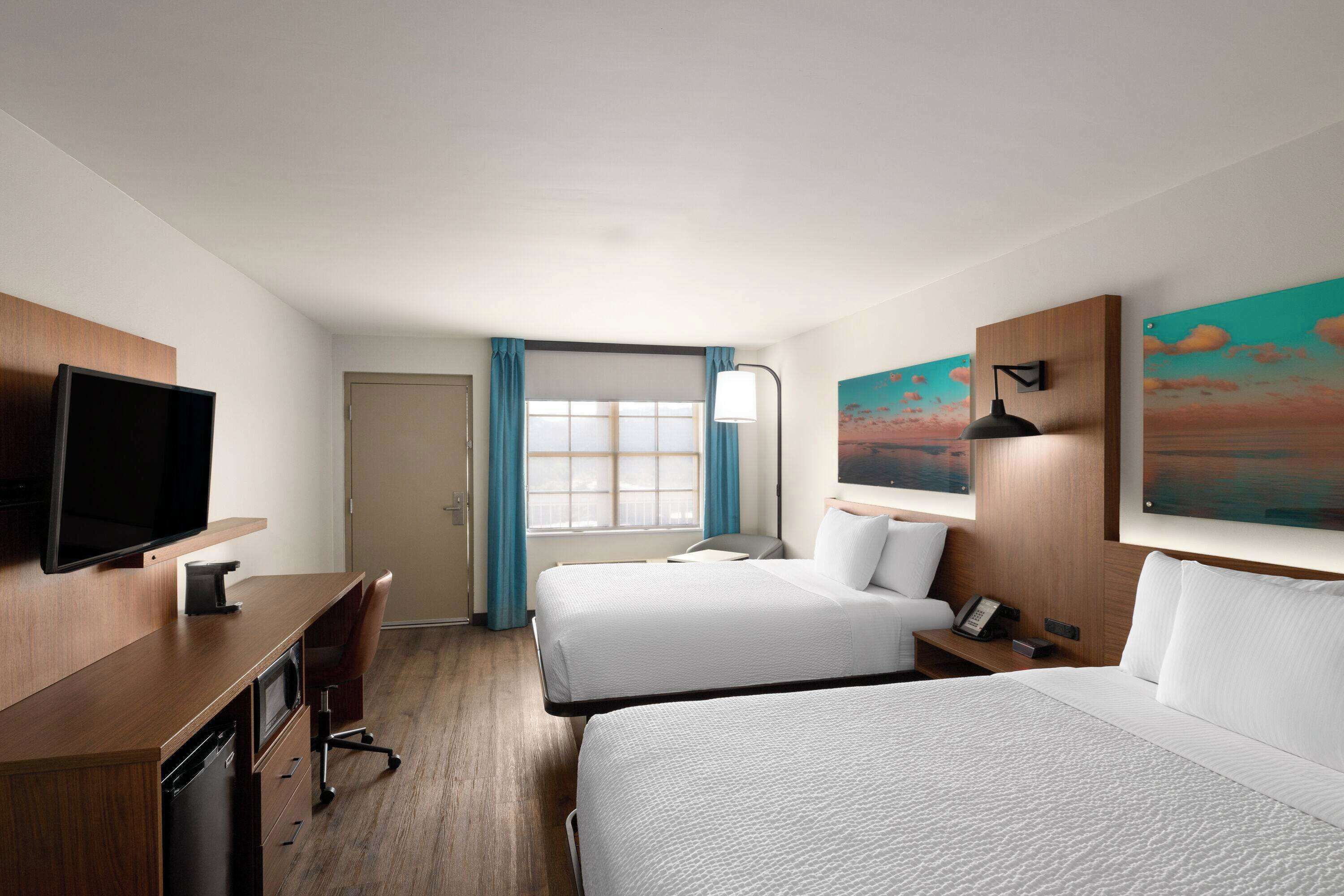 Quarto de hotel com duas camas, uma escrivaninha, televisão e janela com cortinas.