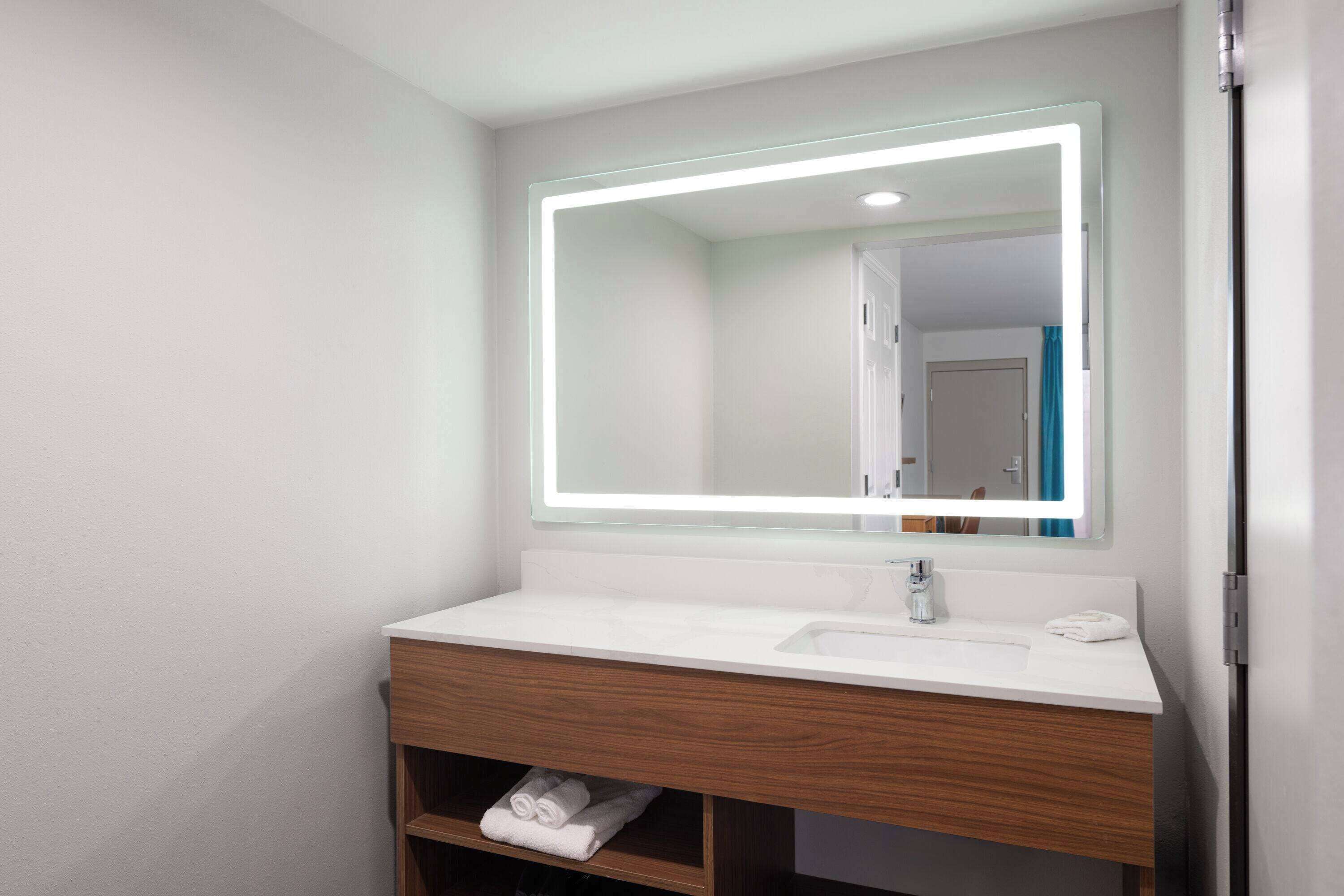 Un baño moderno con lavamanos blanco, tocador de madera y espejo con marco iluminado.