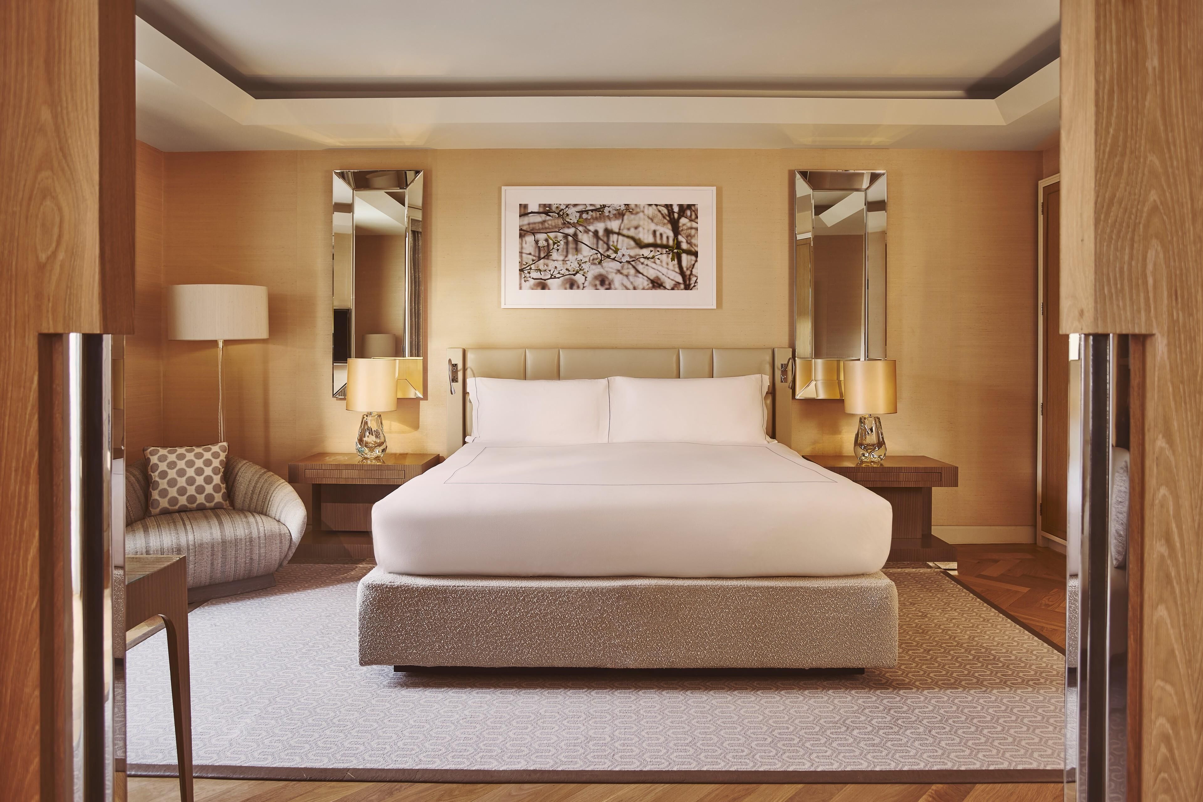 Habitación de hotel con una cama grande, dos mesitas de noche con lámparas, un sofá, un cuadro enmarcado en la pared y un espejo.