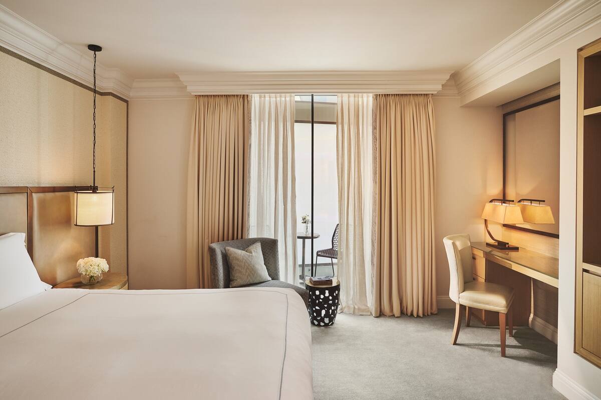 A hotel room with a large bed, a desk, a chair, a lamp, and a view through the window.