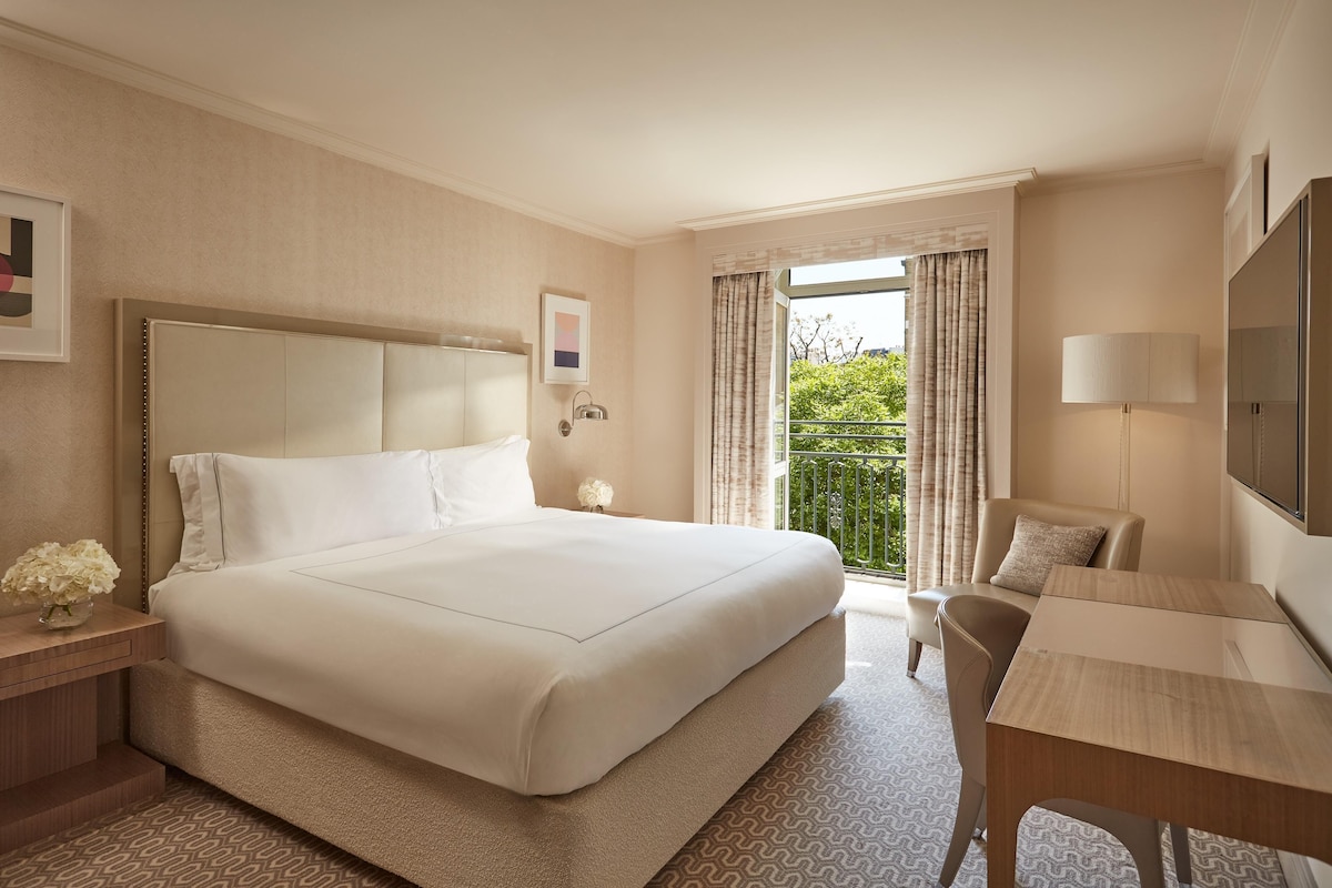 A hotel room with a large bed, a desk, a chair, a TV, and a balcony with curtains.