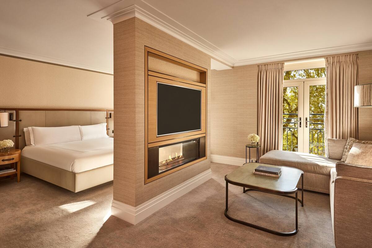 A hotel room with a large bed, a flat-screen TV, a fireplace, a sofa, and a balcony with curtains.