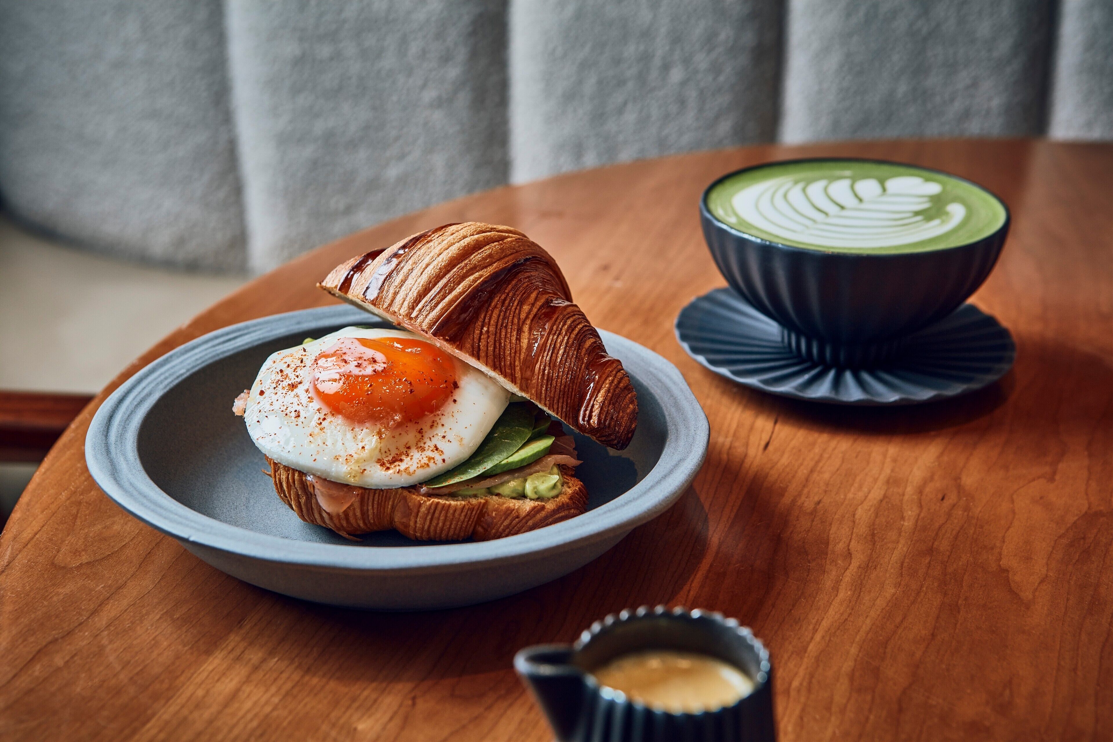 A croissant sandwich with a poached egg and avocado on a plate, a cup of green tea latte, and a small cup of coffee on a wooden table.