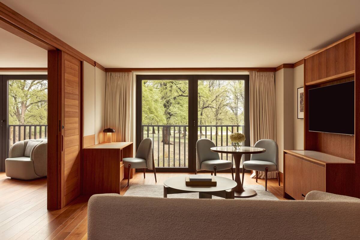 A modern living room with a sofa, two chairs, a round table, a flat-screen TV, and a balcony with a view of trees.
