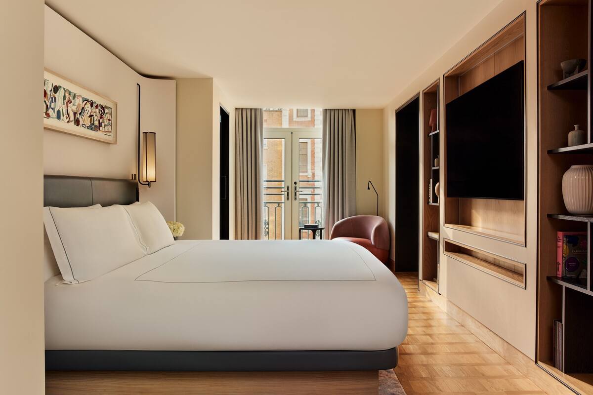 A hotel room with a bed, a TV, a bookshelf, and a view of the outside.