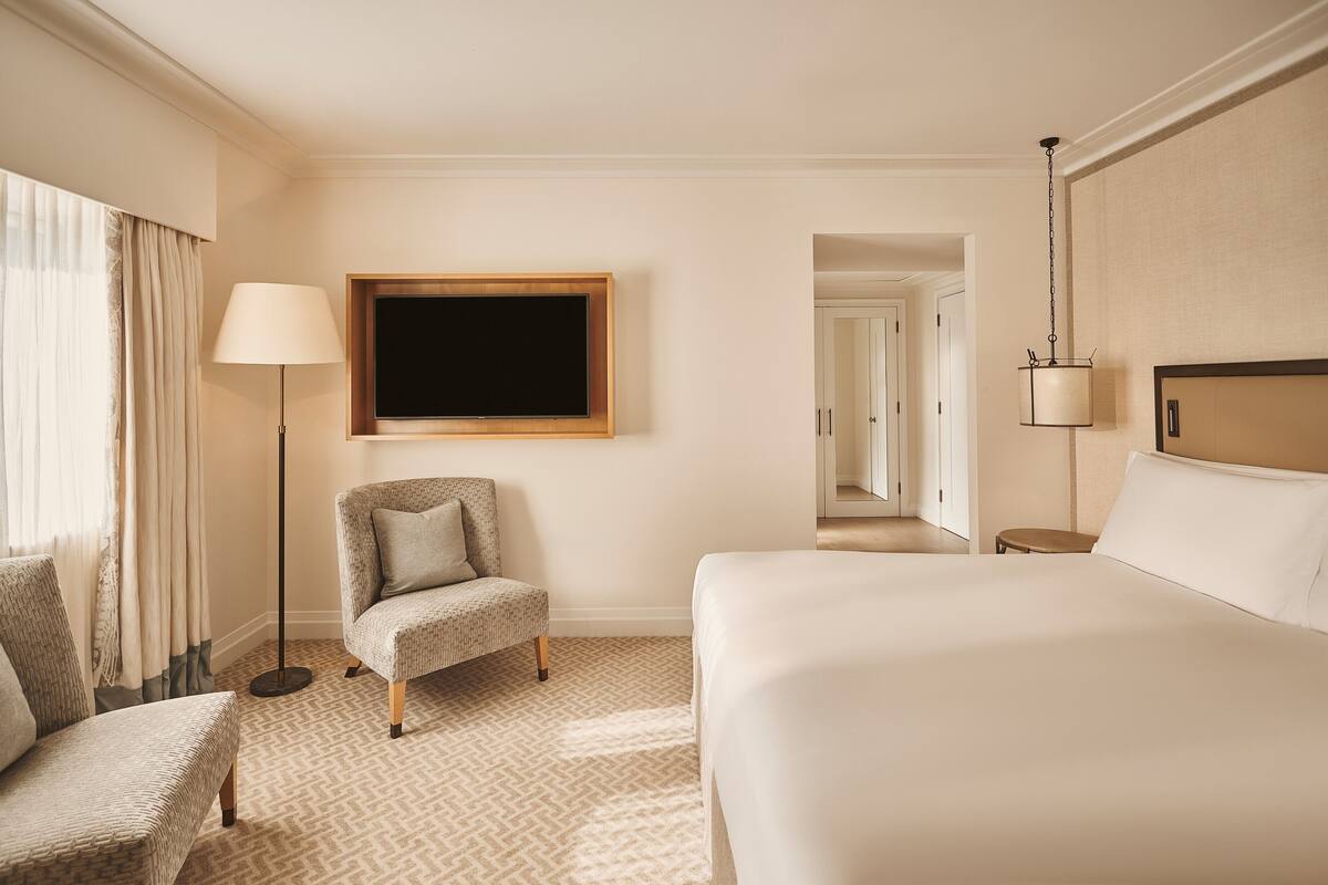 A hotel room with a large bed, a chair, a floor lamp, and a television mounted on the wall.