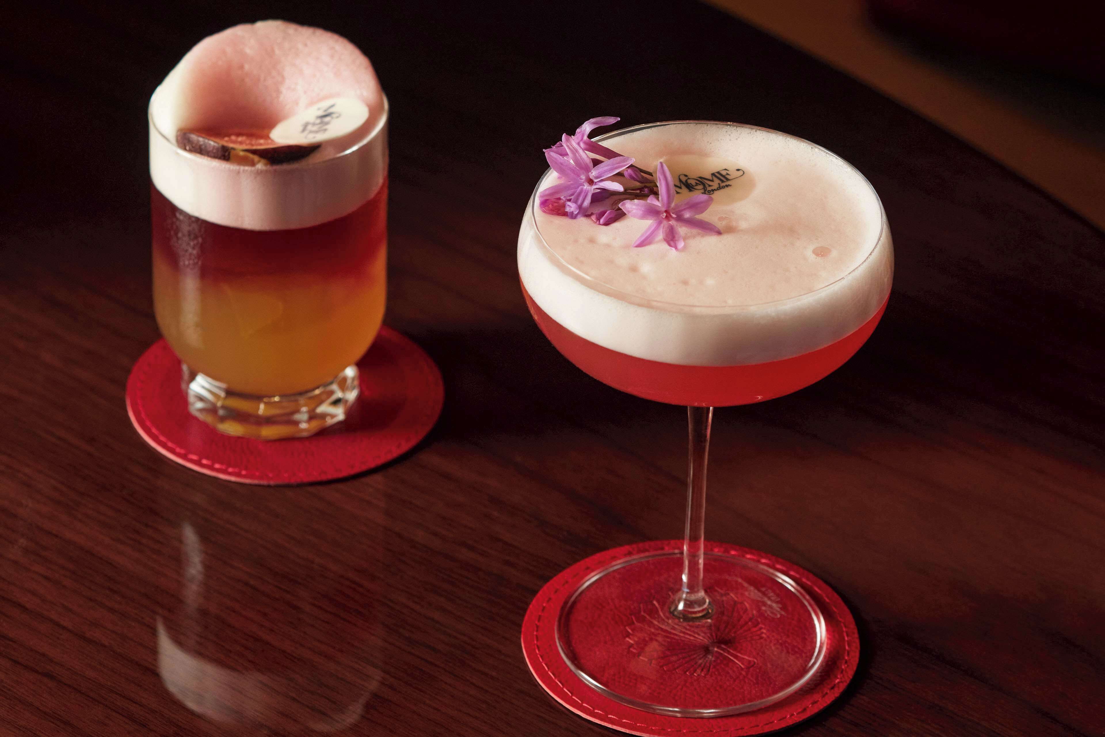 Deux cocktails servis sur des sous-verres rouges, l’un avec une boisson à plusieurs couches et l’autre agrémenté d’une garniture.