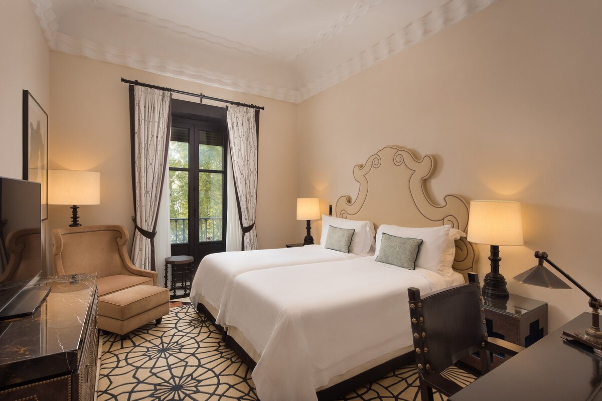 A hotel room with a large bed, two bedside lamps, a chair, a desk, and a patterned floor.