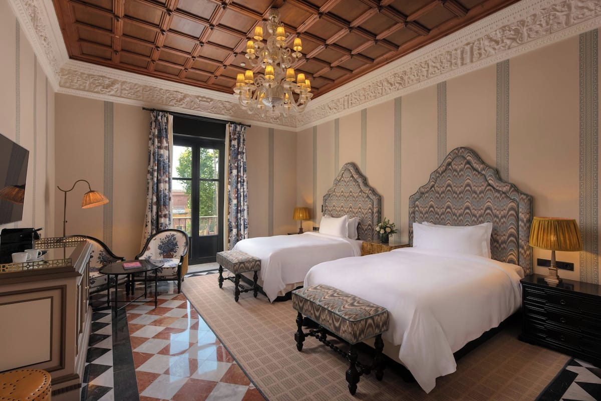 A hotel room with two beds, a chandelier, a desk with a lamp, and a view of the outdoors.