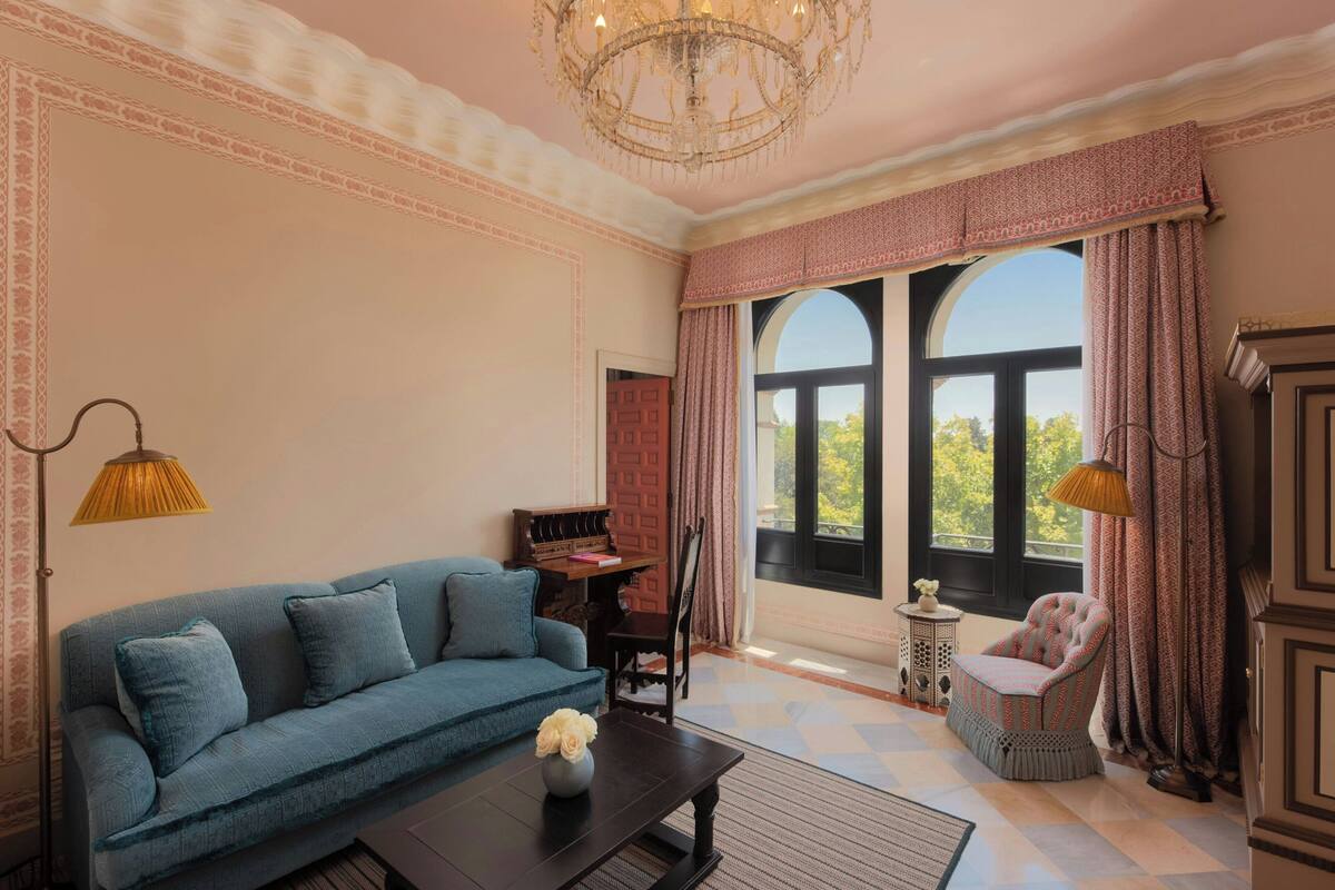 A room with a blue sofa, a small table with a vase, a wooden chair, and a large window with curtains.