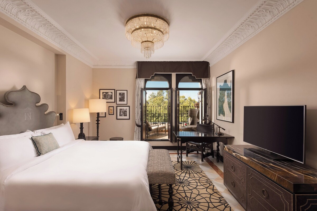 Een hotelkamer met een groot bed, een televisie, een bureau met stoel en een balkon met uitzicht op bomen.