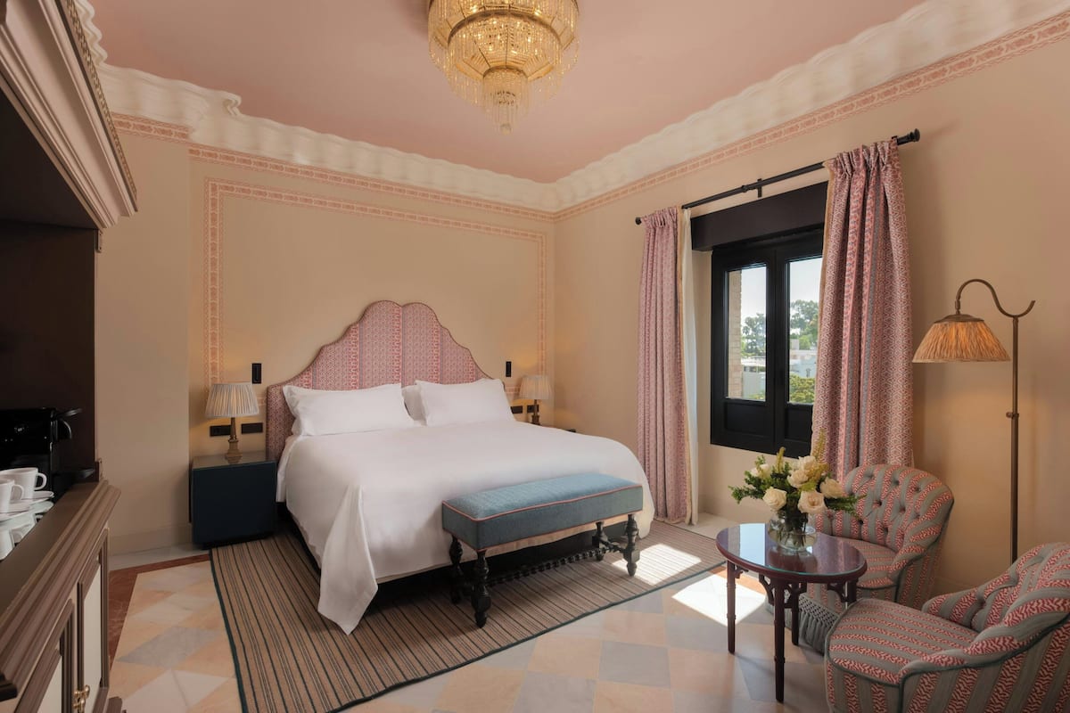 A hotel room with a large bed, a bedside table with a lamp, a chair, a small table with a vase of flowers, and a window with curtains.