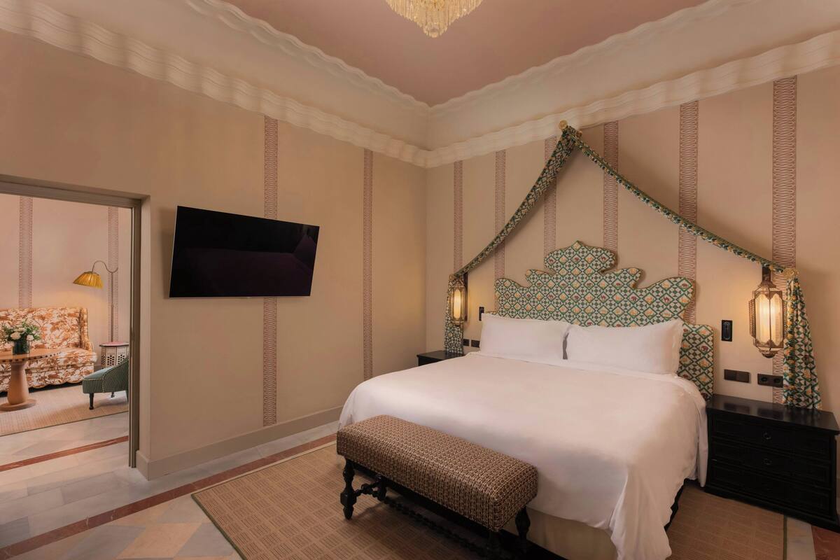 A neatly arranged bedroom with a large bed, a bench, a TV mounted on the wall, and a patterned headboard.