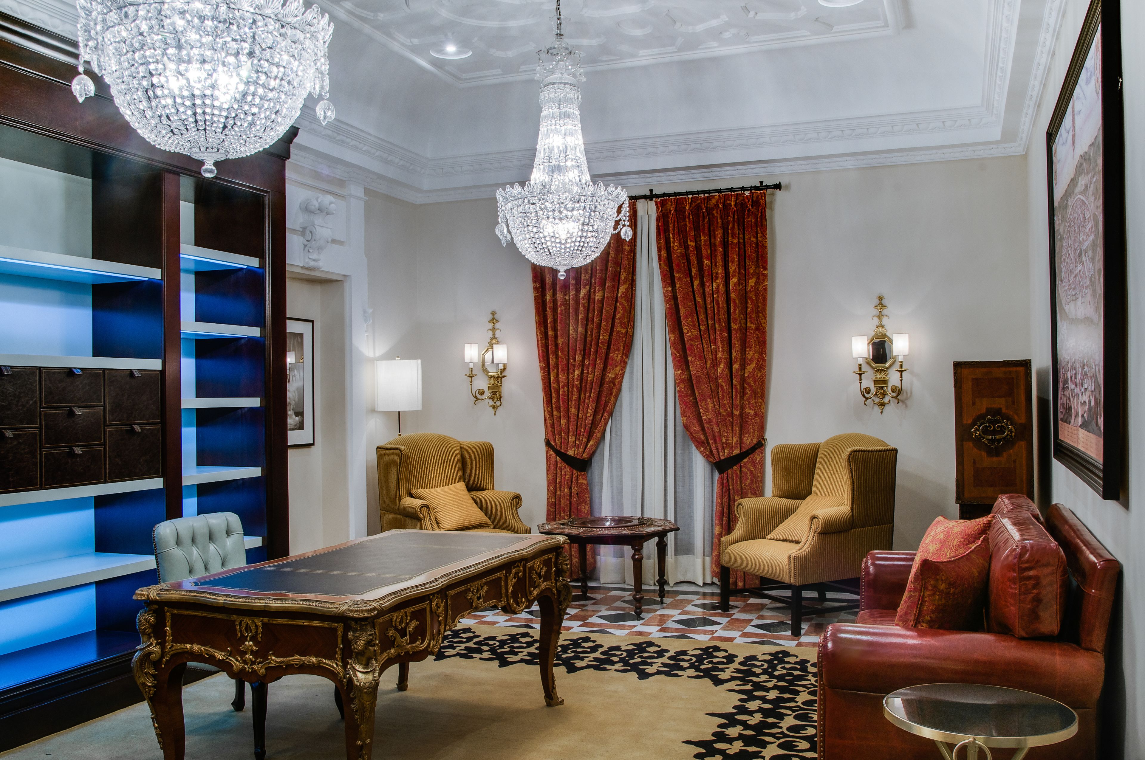 A luxurious room with a large wooden desk, two armchairs, a red sofa, a chandelier, and a large bookshelf.