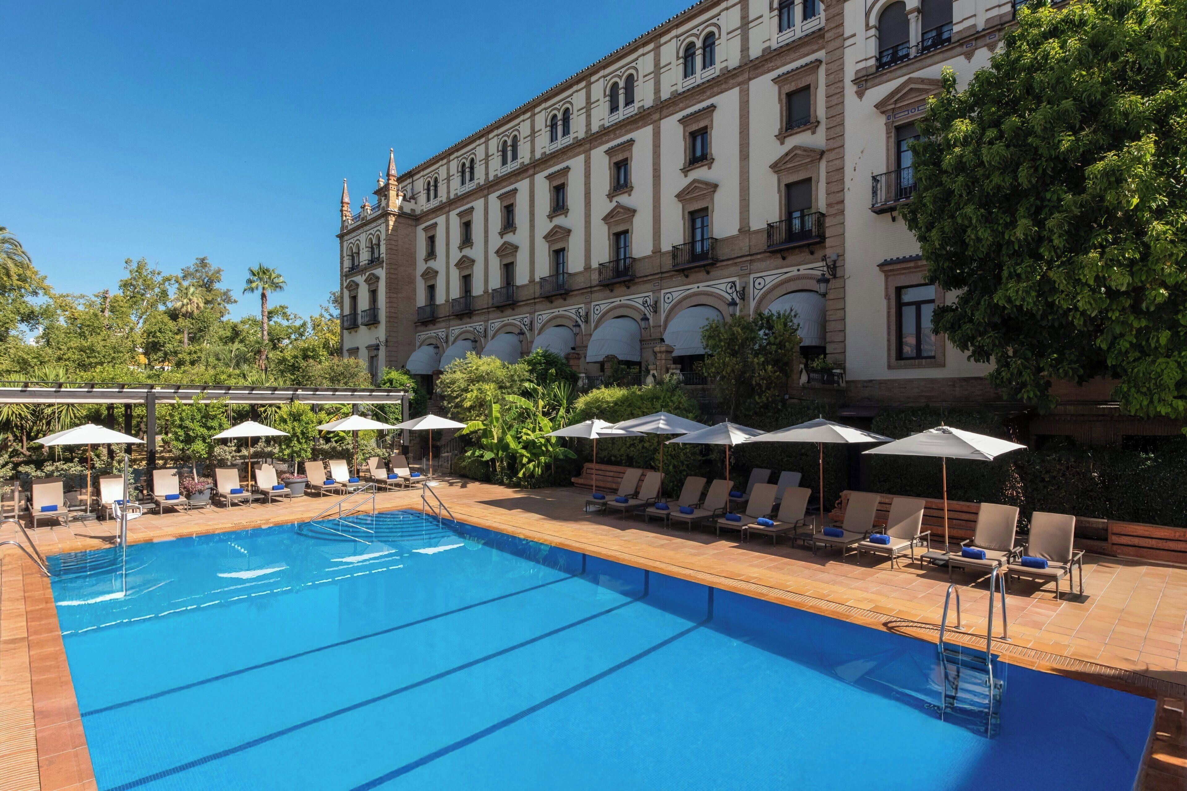 Una piscina con sillas de descanso y sombrillas, rodeada por un edificio de arquitectura clásica.