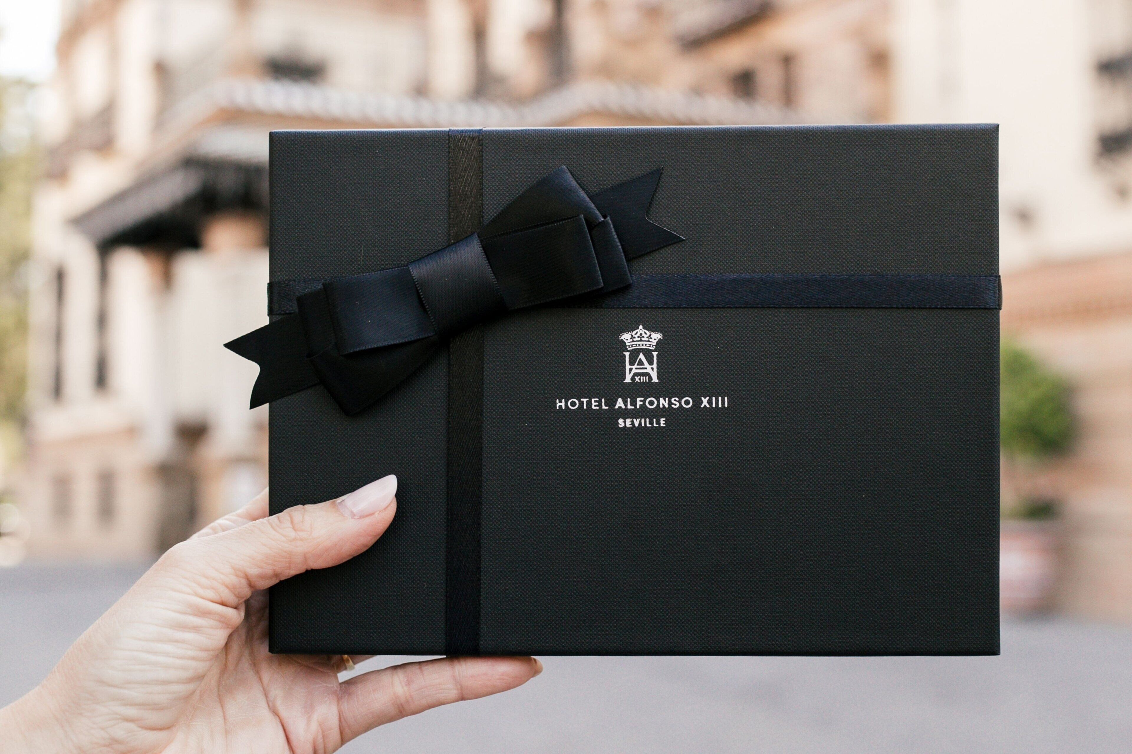 Una caja de regalo negra con un lazo y cinta negros, etiquetada como "HOTEL ALFONSO XIII SEVILLE".