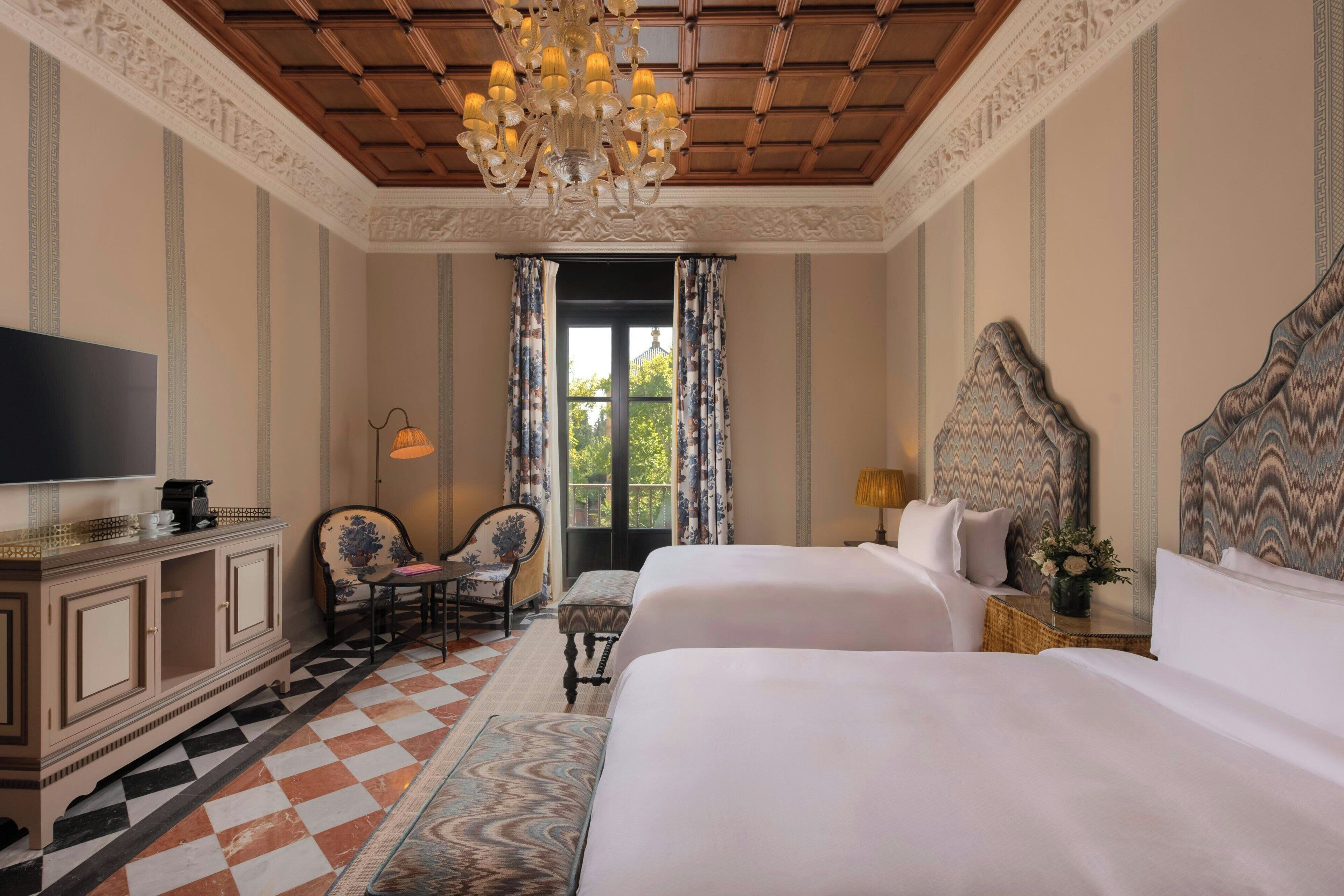 A hotel room with two beds, a television, a desk with a chair, a chandelier, and a large window with curtains.