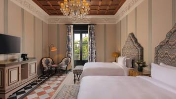 A hotel room with two beds, a television, a desk with a chair, a chandelier, and a large window with curtains.