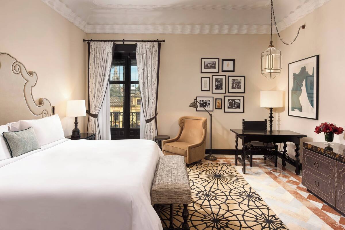 A hotel room with a large bed, a chair, a desk, and a dresser. There are pictures on the wall and a window with curtains.