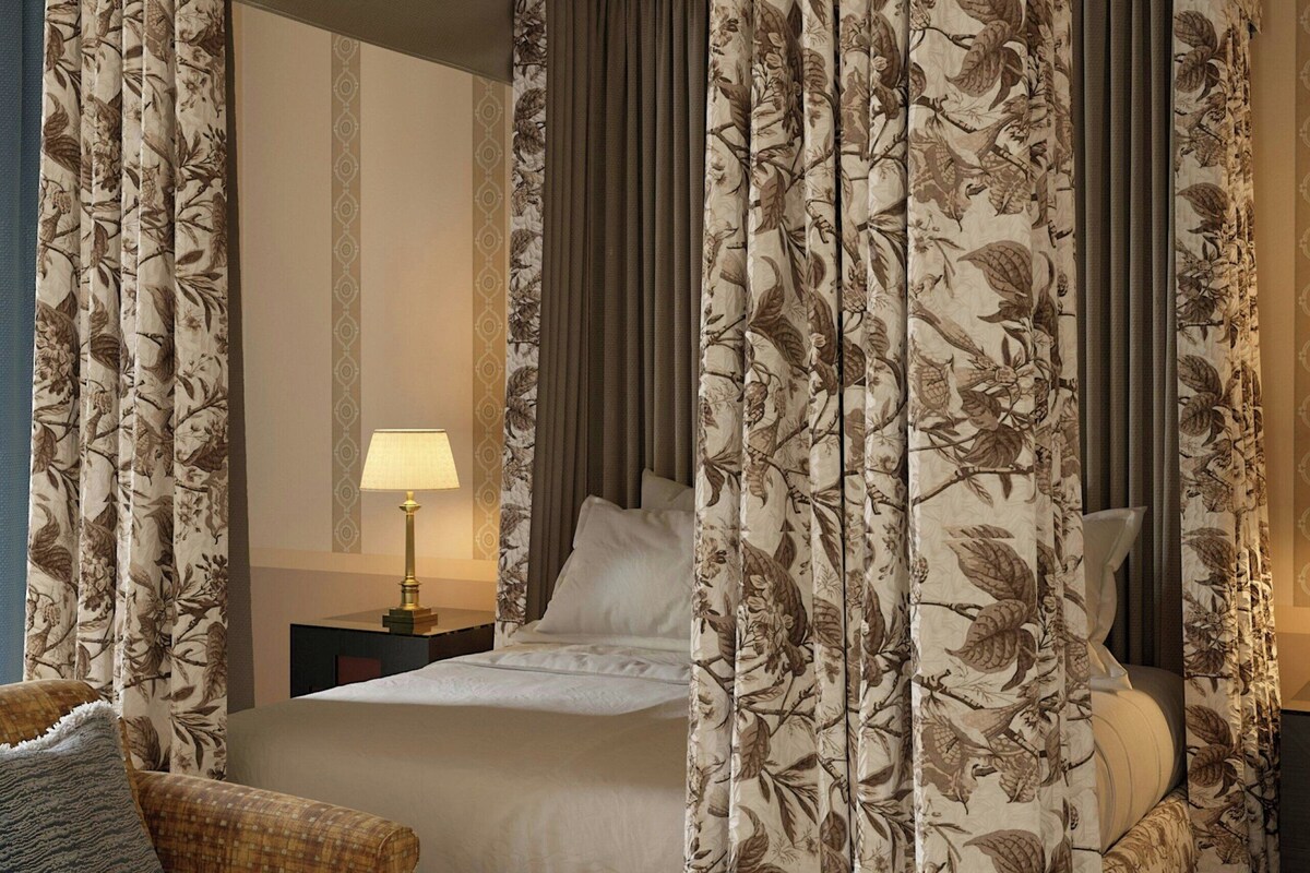 Uma cama com dossel e cortinas florais, uma mesa de cabeceira com abajur e um sofá com almofada.