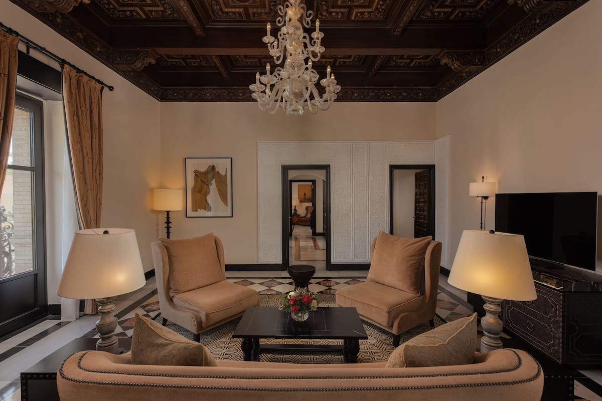 A hotel room with a large sofa, two armchairs, a coffee table with a vase of flowers, a TV, and a chandelier.