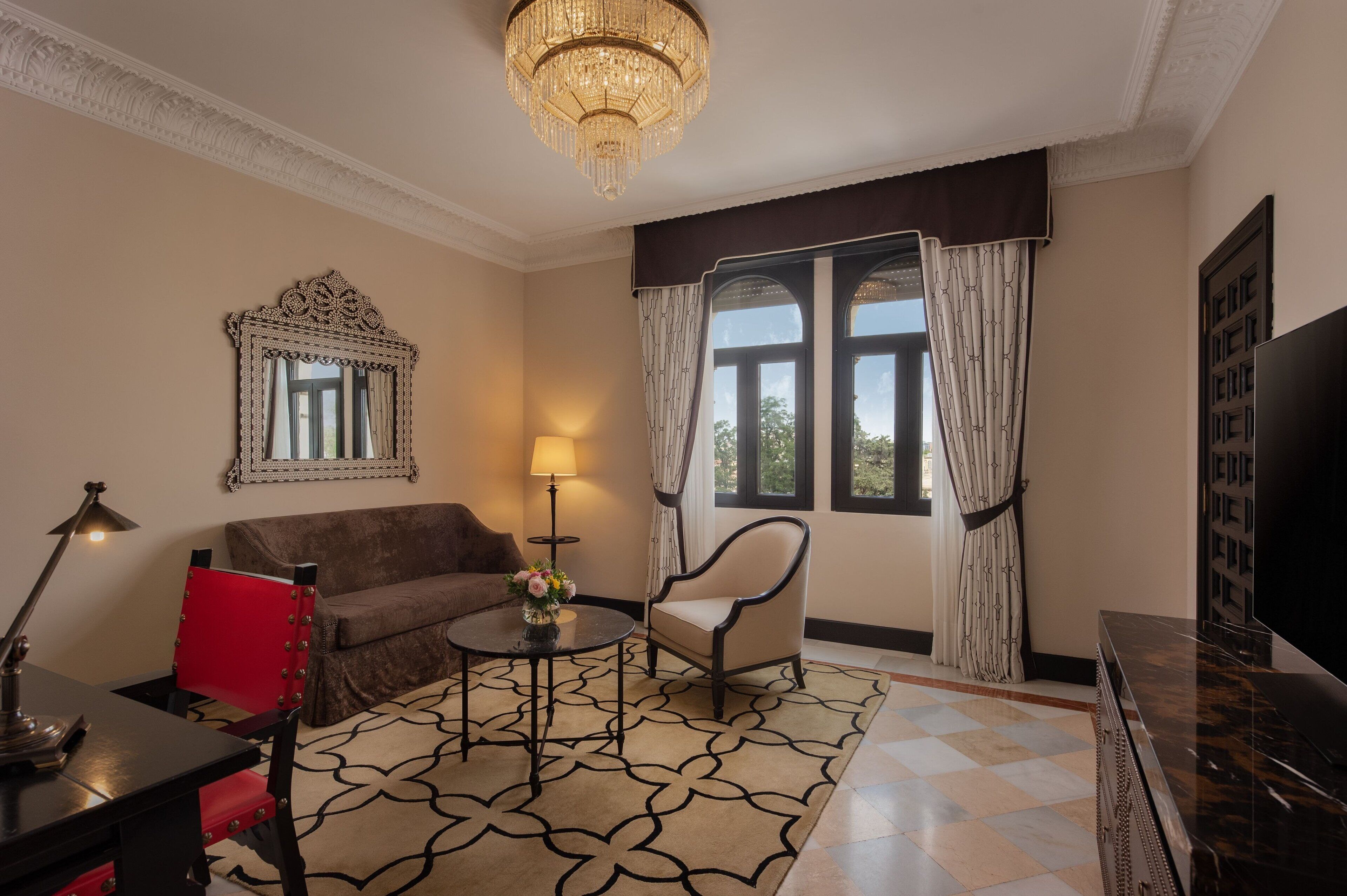 Una habitación con candelabro, sofá, sillón, mesa con lámpara, televisor y ventana con cortinas.