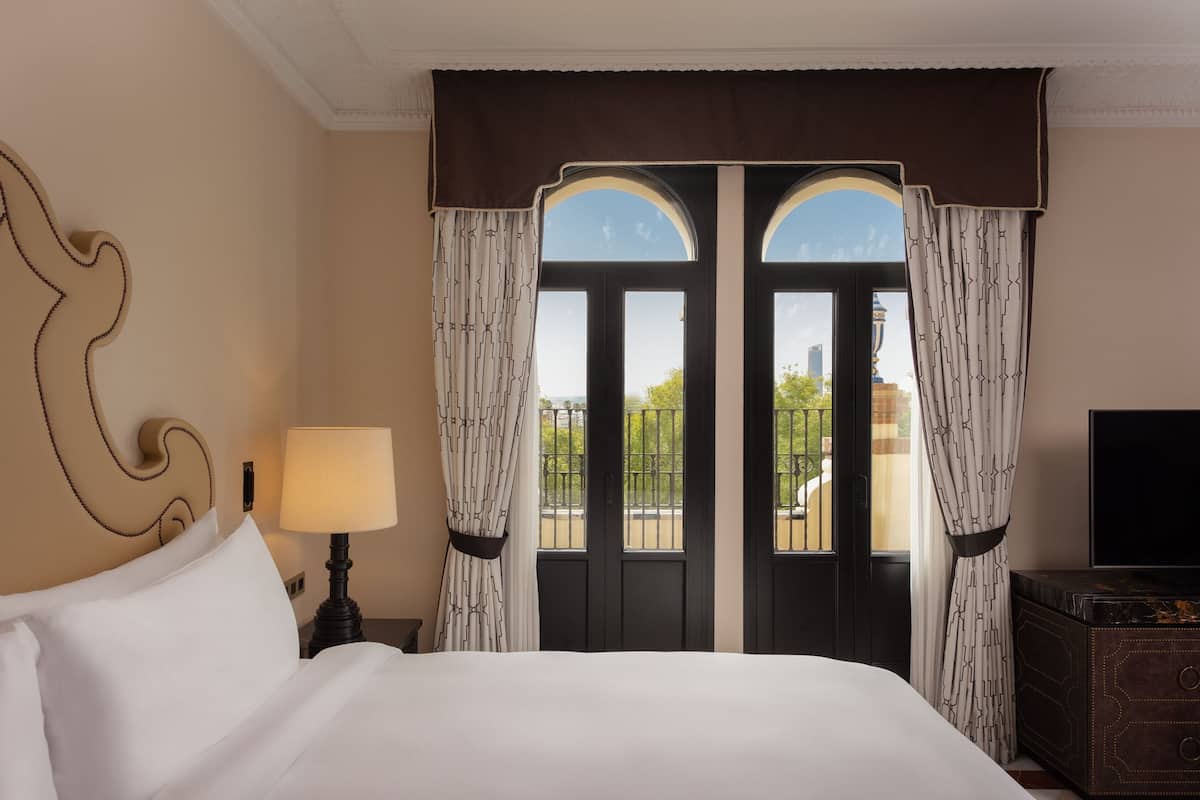 A hotel room with a bed, a nightstand with a lamp, a TV, and a view of the outside through a double door.