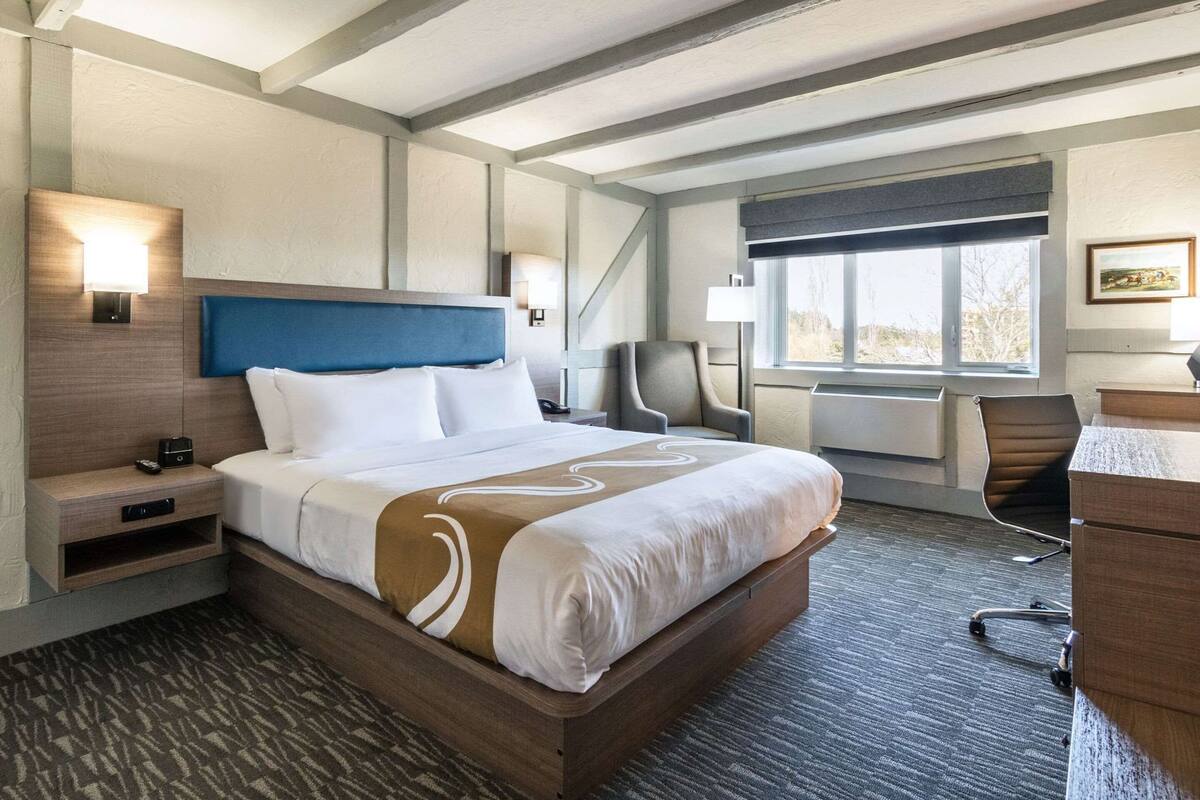 A hotel room with a large bed, a desk, a chair, a nightstand, and a window with a view.