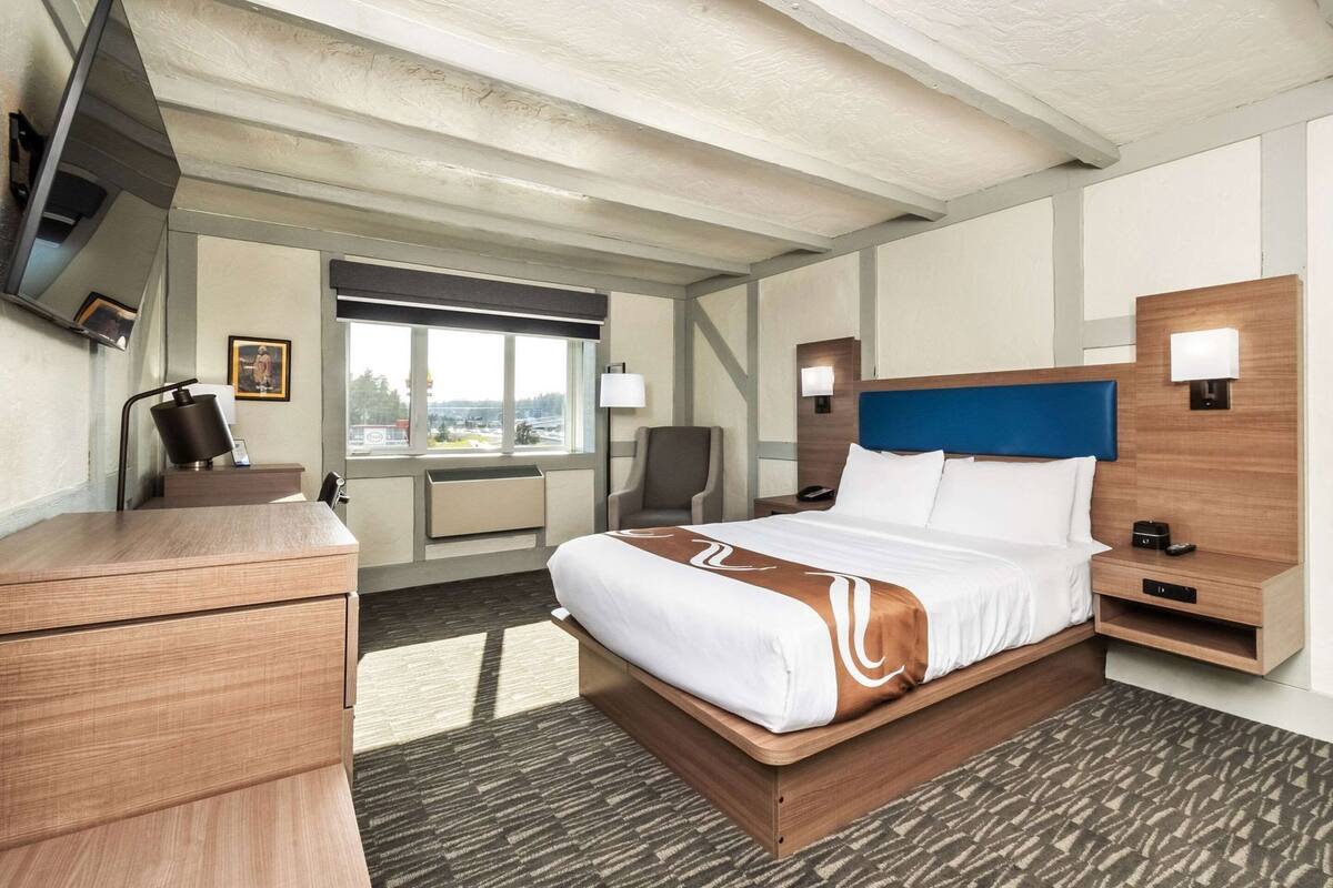 A hotel room with a bed, desk, chair, bedside tables, lamp, and a window with a view.