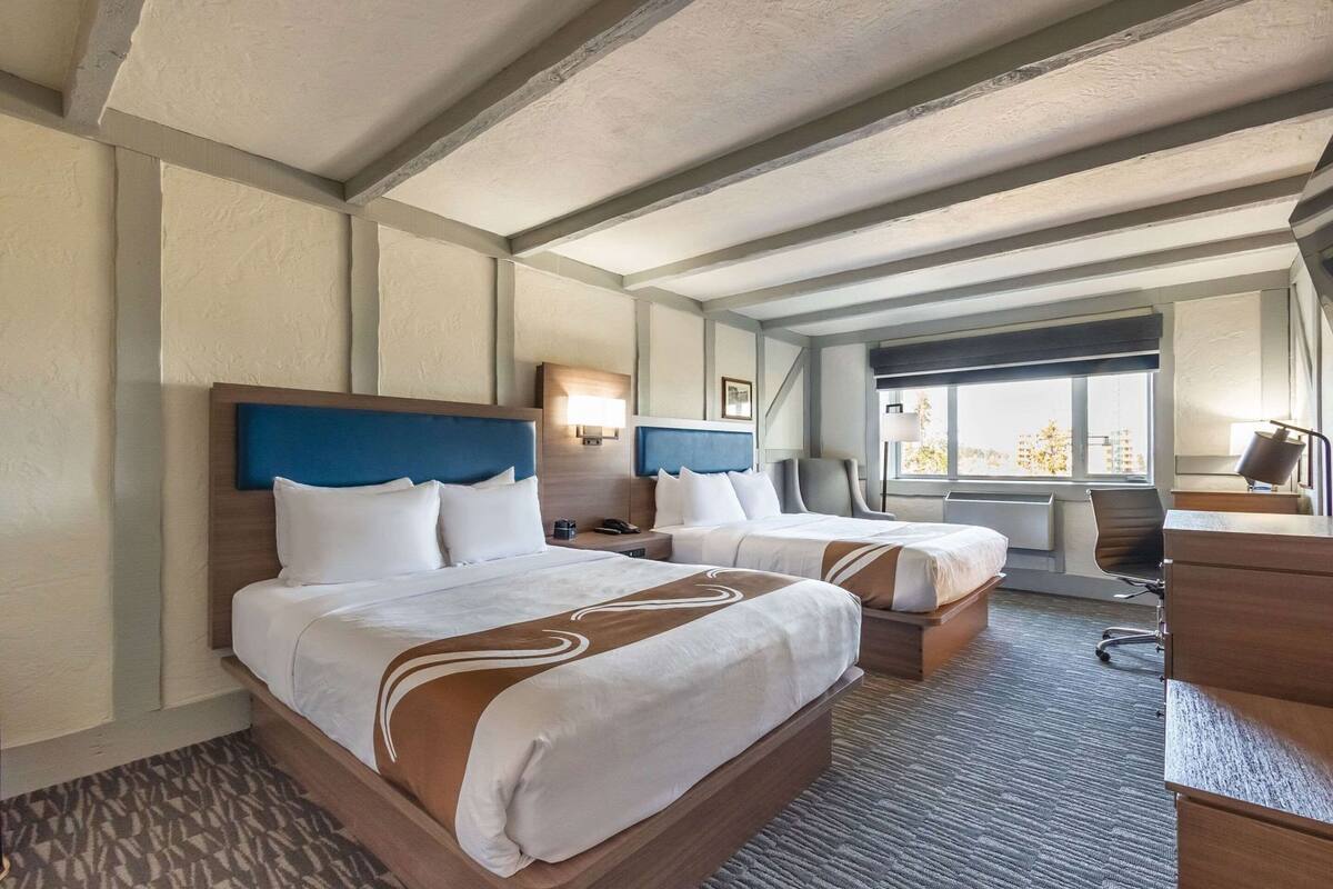 A hotel room with two beds, a desk, a chair, a lamp, and a view of the outside.