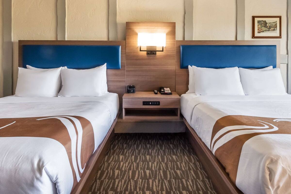 A hotel room with two beds, a nightstand with a lamp, and a small table.